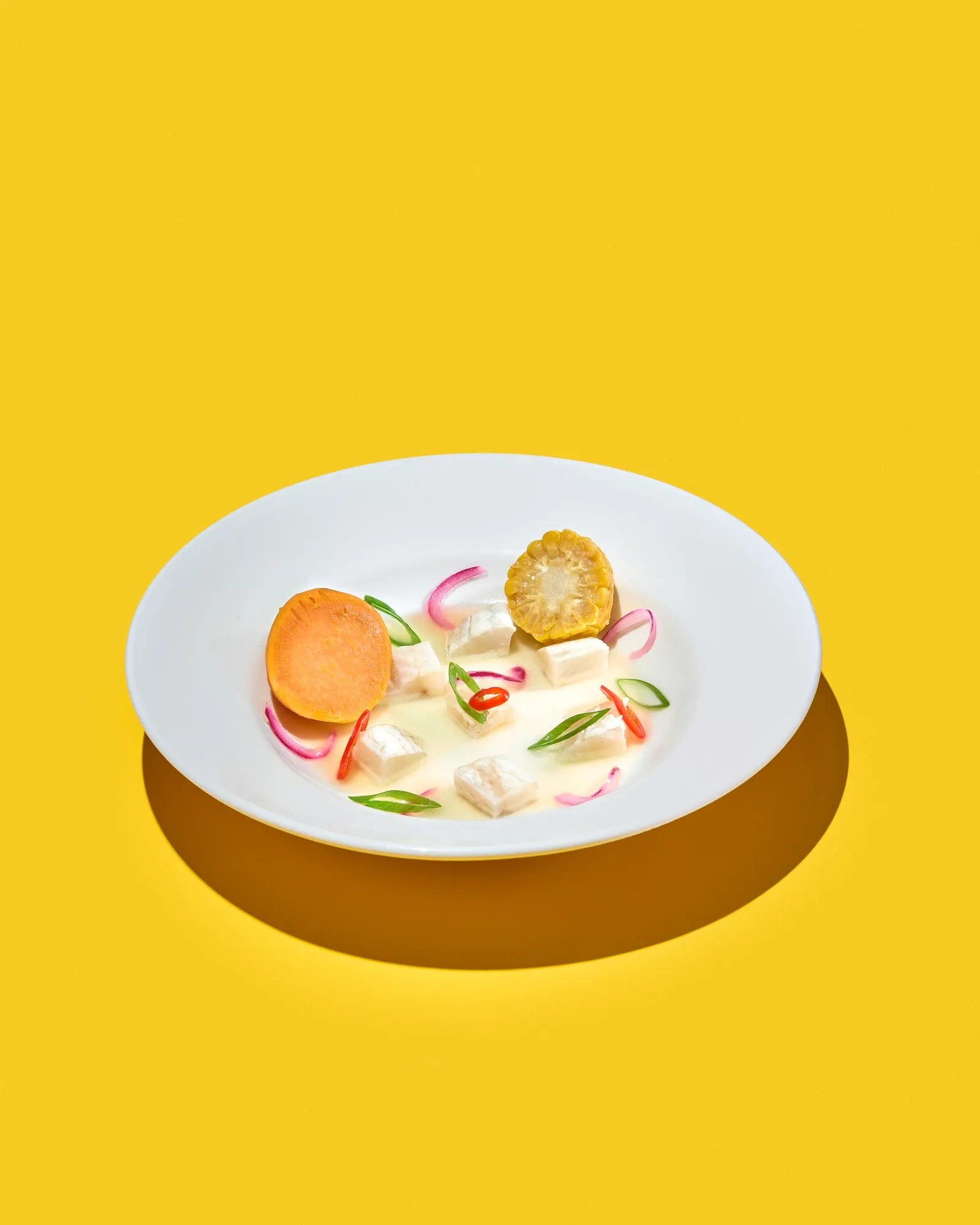 Cubes of white fish in a milky broth with half a sweet potato, a round of corn on the cob, pink pickled onion, chilli and spring onion, in a wide white bowl against flat yellow, shot for the cookbook 17 Perspektiven