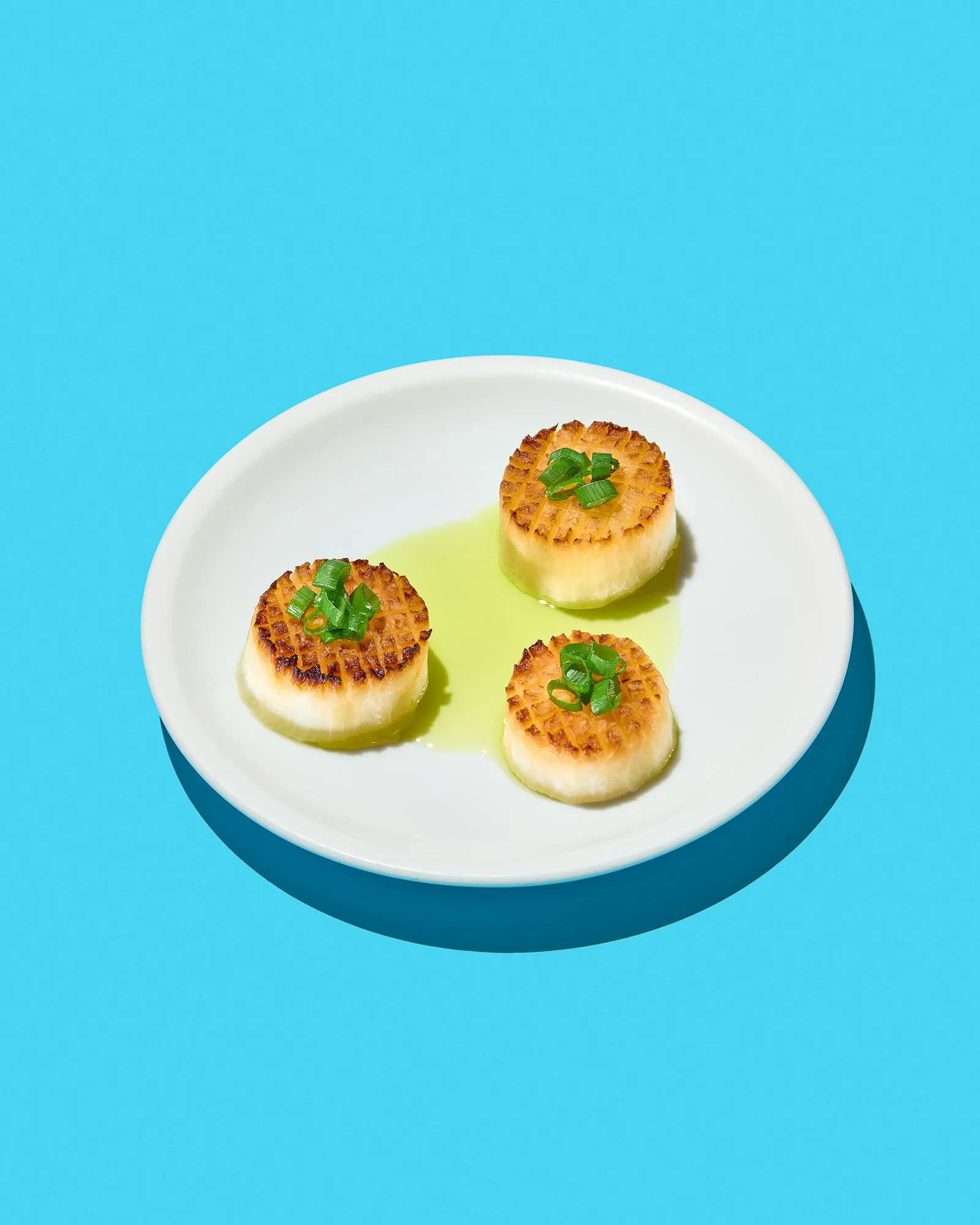 Three seared rounds of daikon with crosshatched golden tops on a white plate, each crowned with spring onion and sitting in a pool of bright green oil, against flat cyan, shot for the cookbook 17 Perspektiven