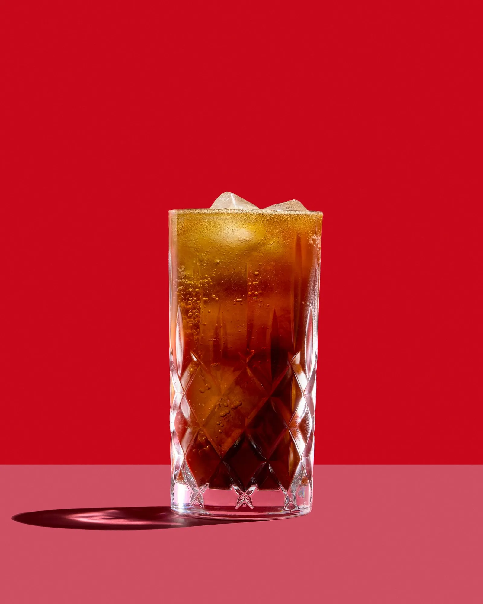 A dark amber sparkling drink over ice in a cut crystal highball, bubbles clinging to the inside of the glass, on a pink ledge against red with a hard shadow, shot for the cookbook 17 Perspektiven