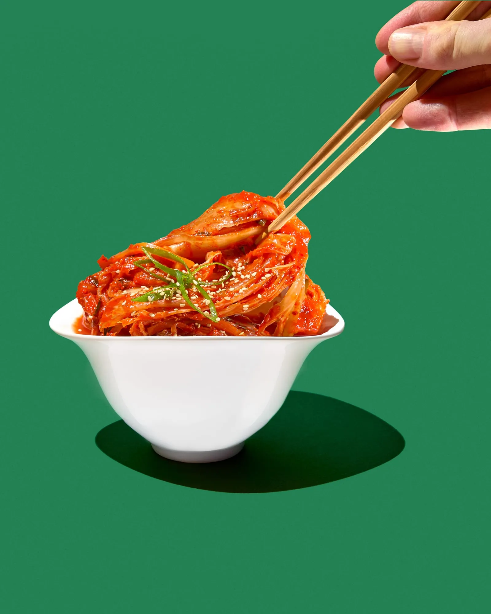 Kimchi piled high in a flared white bowl, a hand lifting a strand of it on wooden chopsticks, sesame and spring onion scattered over the top, against flat green with a hard shadow, shot for the cookbook 17 Perspektiven