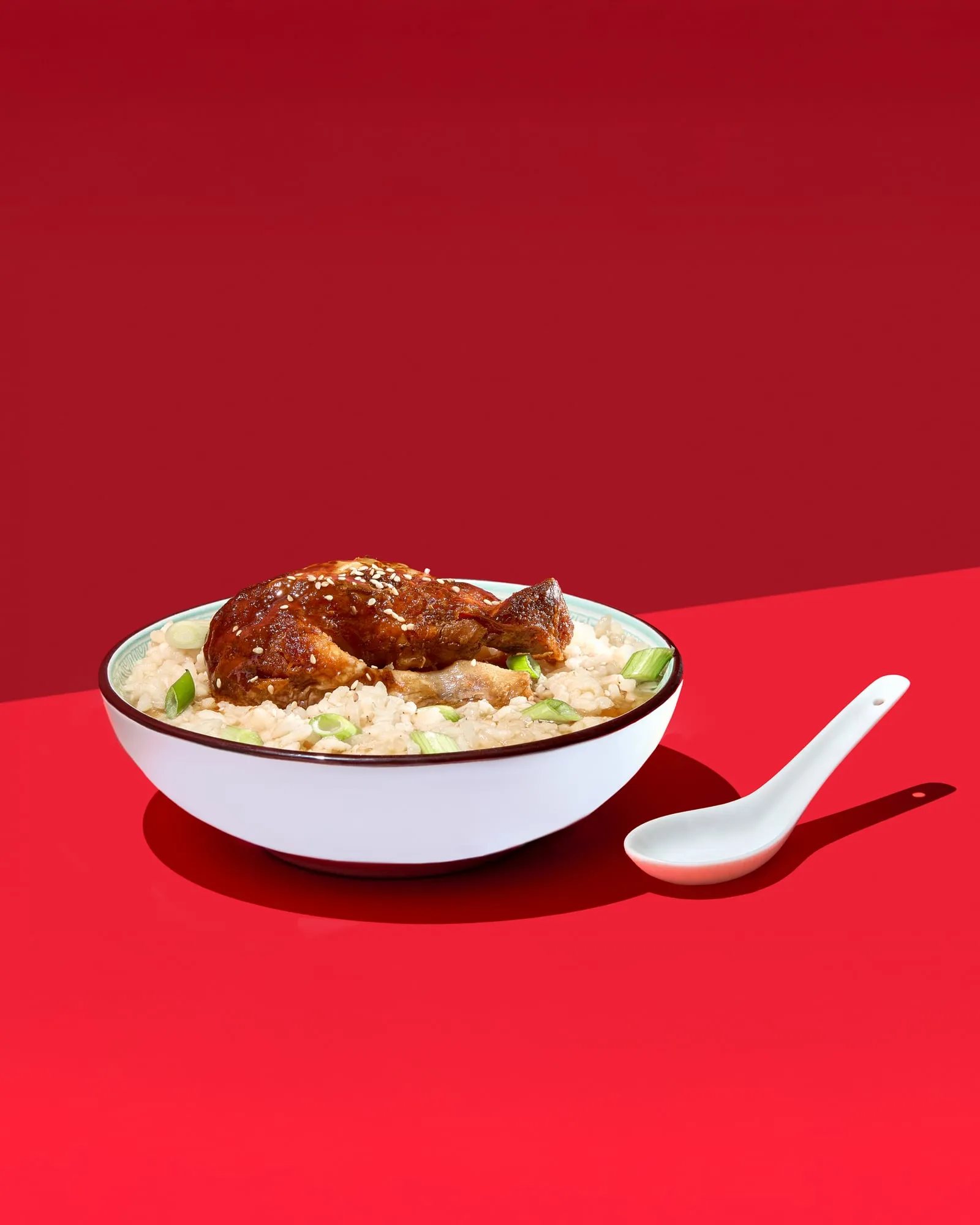 A glossy sesame glazed piece of braised meat on a bowl of rice in broth with spring onion, a white ceramic spoon beside it, on two tones of red under hard sunlight, shot for the cookbook 17 Perspektiven