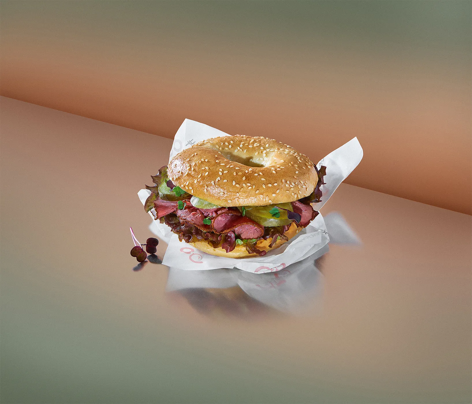 Sesame bagel filled with pastrami, pickles and leaves on its wrapper, peach and green backdrop