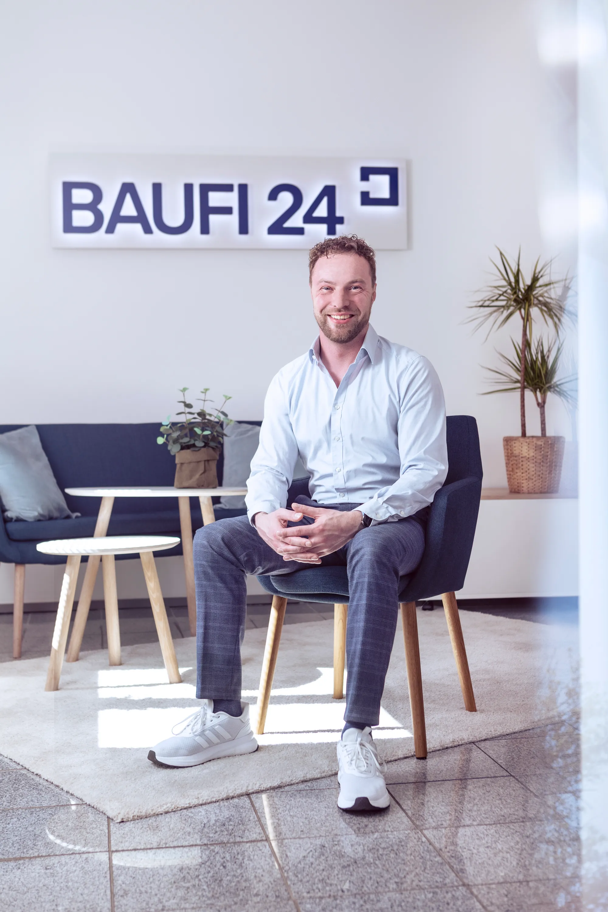 A man in a pale blue shirt and checked trousers sitting forward in a dark armchair beneath the Baufi 24 sign, hands loosely clasped, the office lounge behind him in low sun with a sofa, a low round table and a potted dracaena