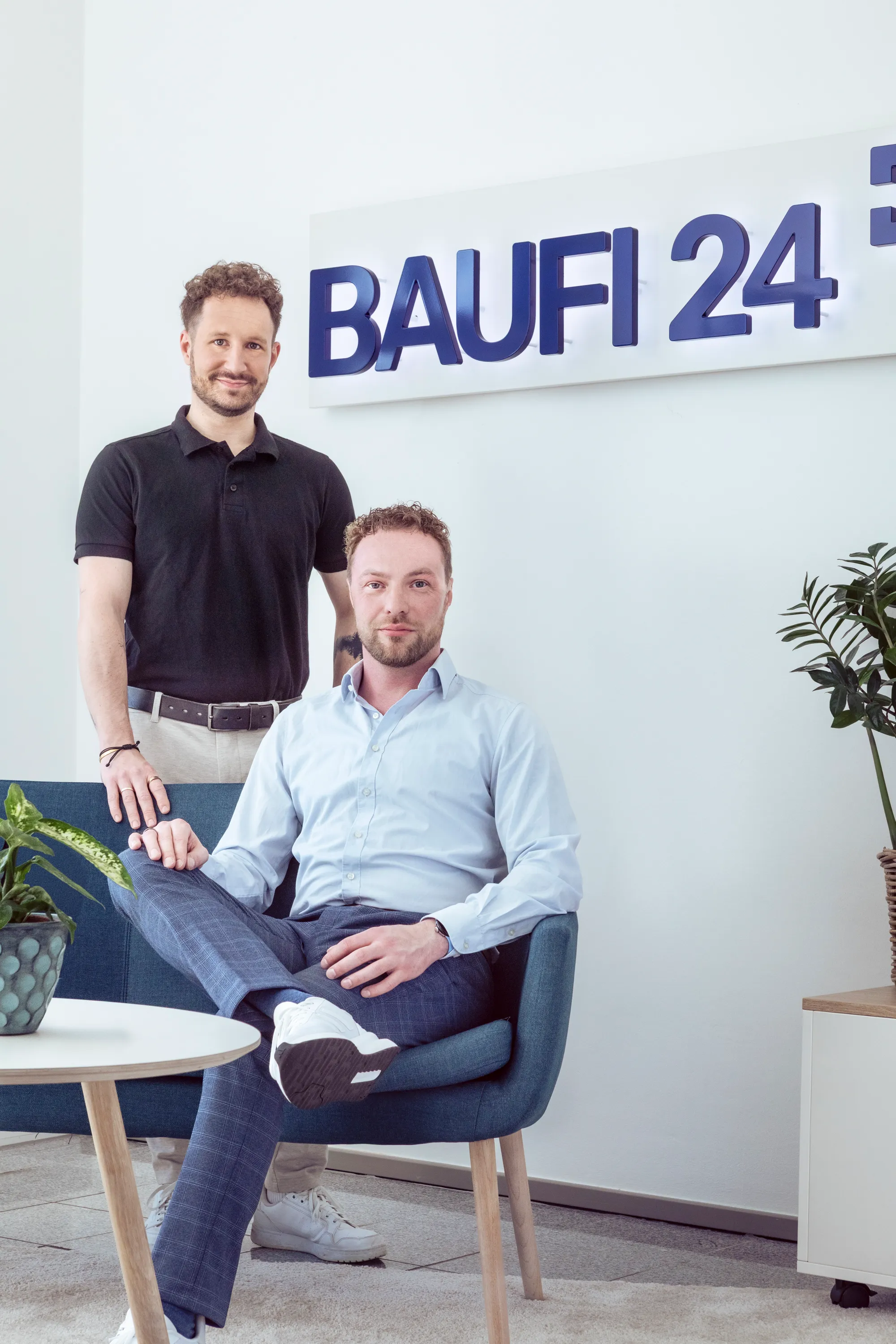 Two men photographed beneath the Baufi 24 sign in their office, one standing behind in a black polo with a hand resting on the armchair, the other seated with his legs crossed, both looking straight down the lens