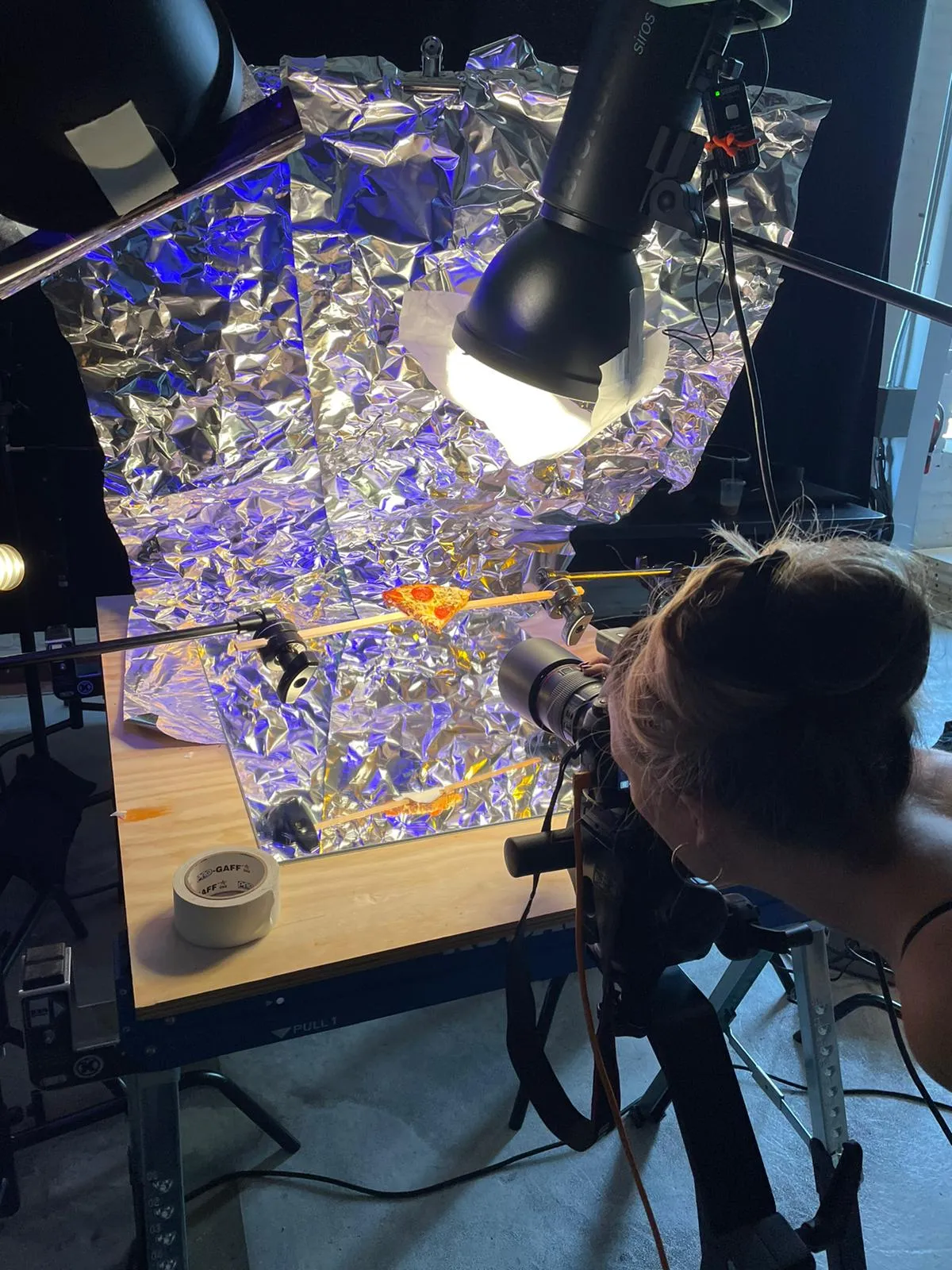 A slice of pepperoni pizza rigged on rods over crumpled foil under blue and orange light, shot from behind the camera