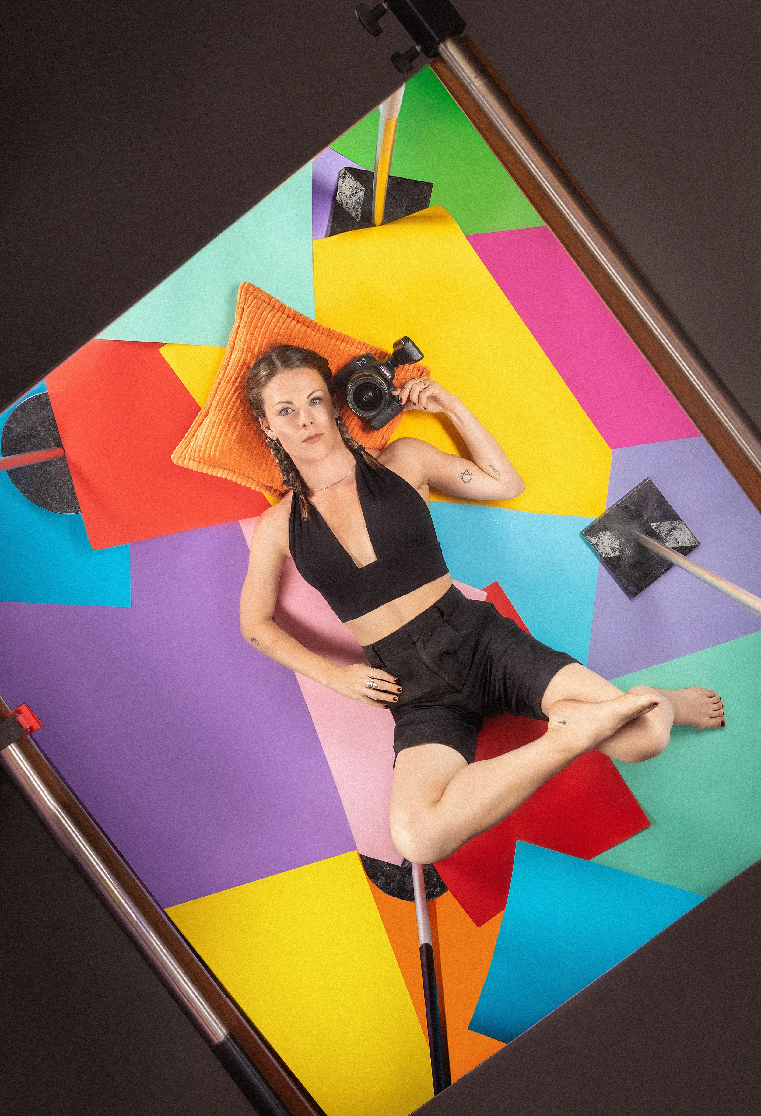 Em Wild lying on her back across a set built from overlapping sheets of coloured paper, camera held up beside her face and looking straight up into the lens, the studio stands and flags cutting across the corners of the frame