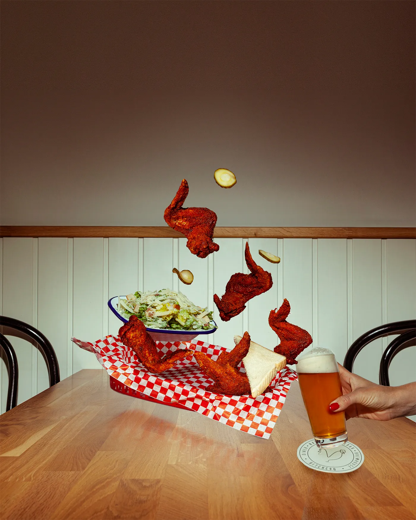 Hot chicken wings mid air above a checked basket with slaw, white bread and a beer on a diner table