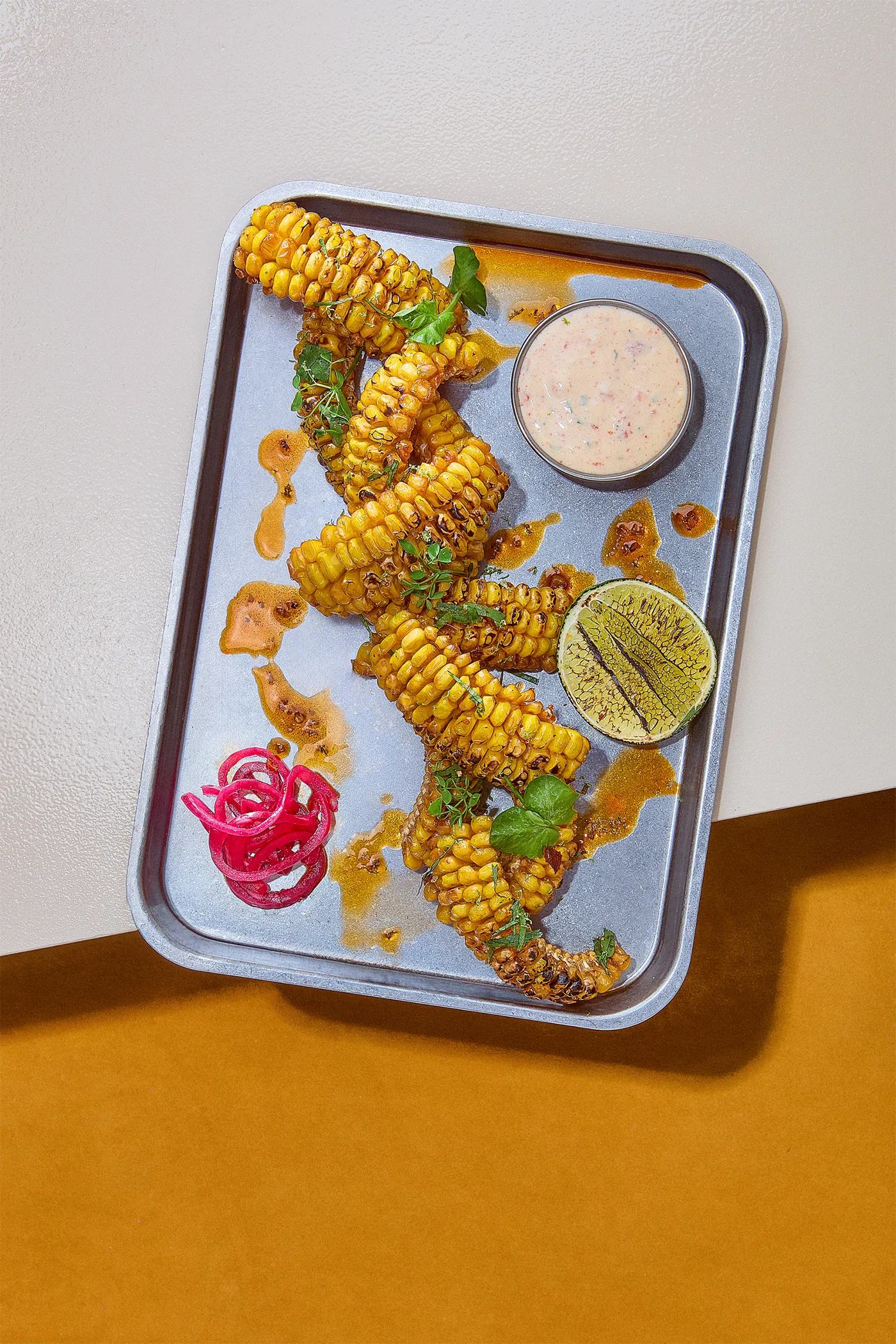 Charred corn ribs on a steel tray with chipotle dip, lime and pickled onion on ochre