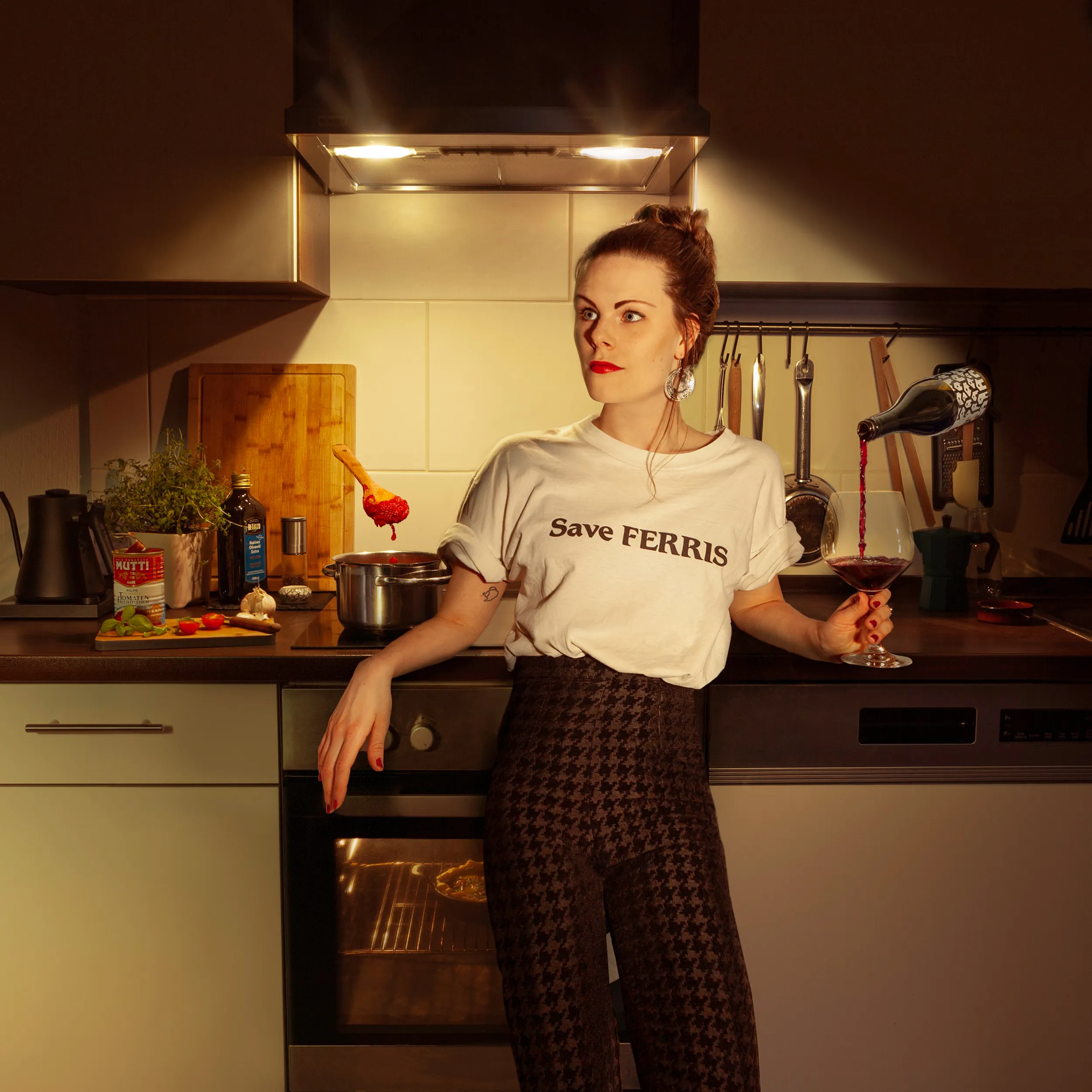 Em Wild in her kitchen in a Save Ferris shirt, red wine pouring itself into her glass and a spoon of tomato sauce hanging mid air