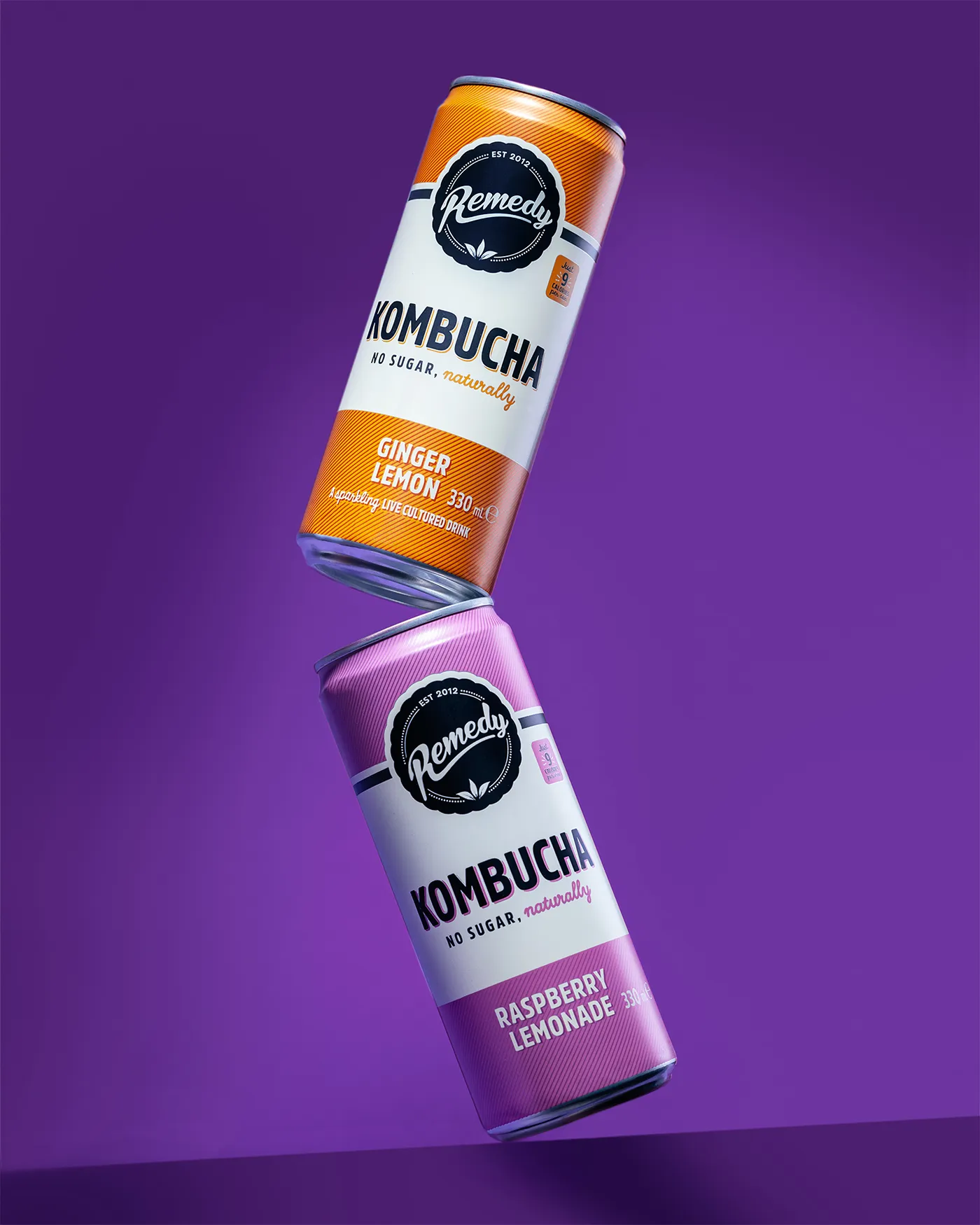 Remedy Kombucha ginger lemon and raspberry lemonade cans balanced one on the other against purple