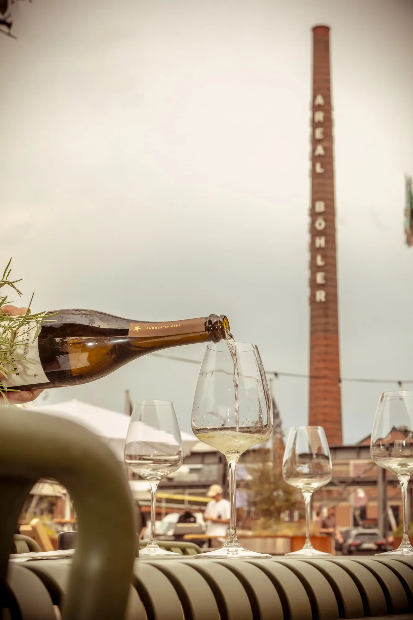 An Andres Mugler bottle pouring white wine into a glass on a terrace table at the Weinfest with Mission Genuss, the Areal Böhler chimney in Düsseldorf rising behind