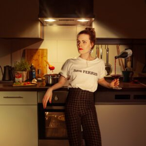 A woman in a "Save FERRIS" t-shirt standing in a kitchen with a levitating spoon of sauce and wine pouring itself into her glass.